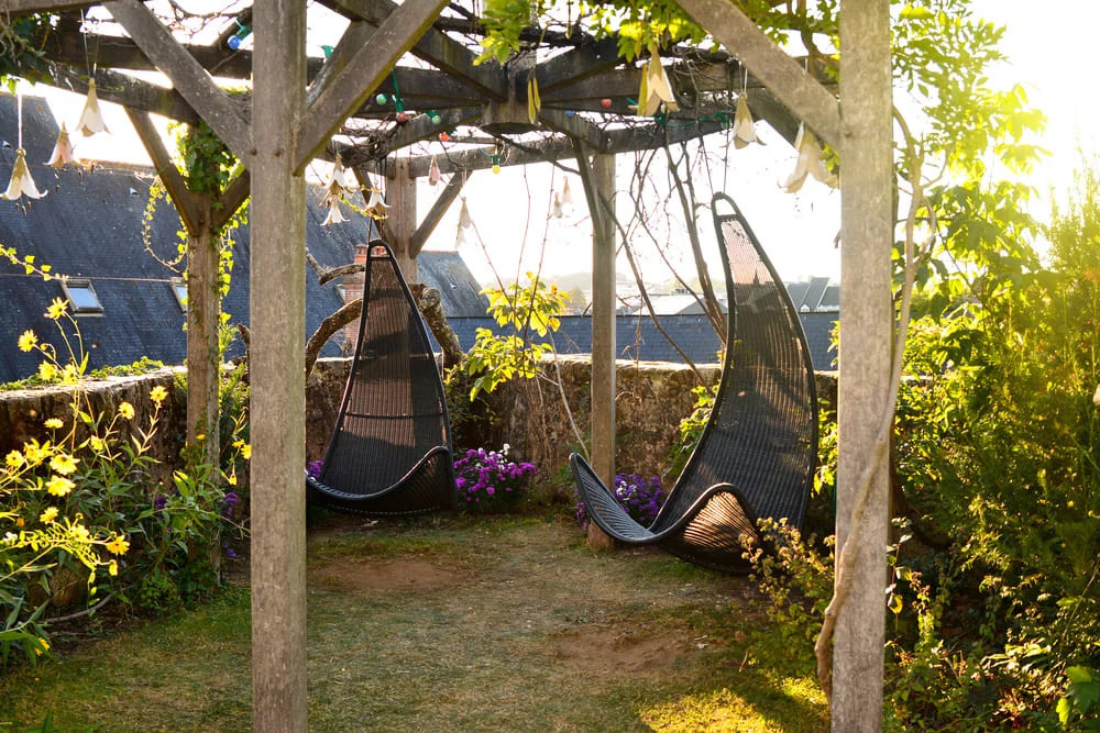Two hanging garden chairs under a wooden pergola in a sunlit garden.
